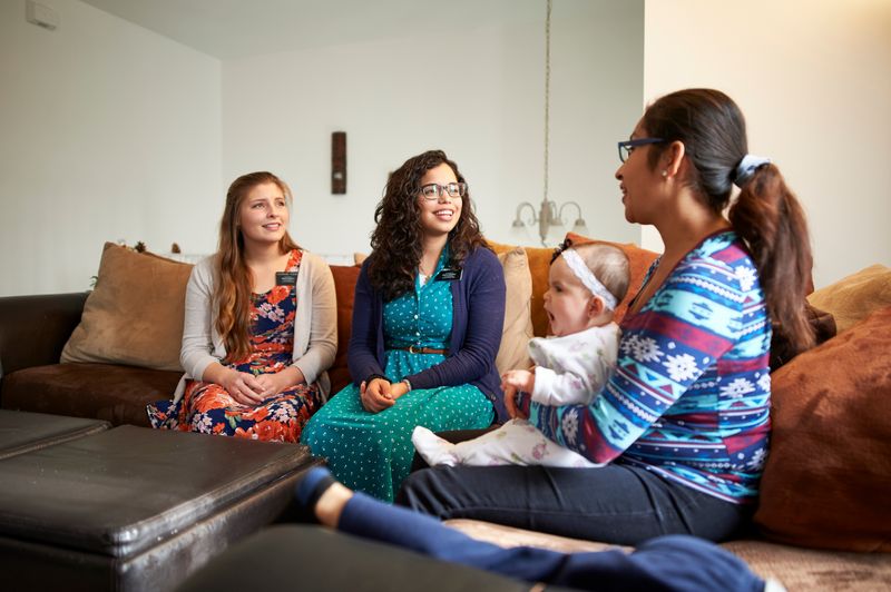 Twee zendelingzusters onderwijzen een jonge moeder en haar zoon in haar woonkamer